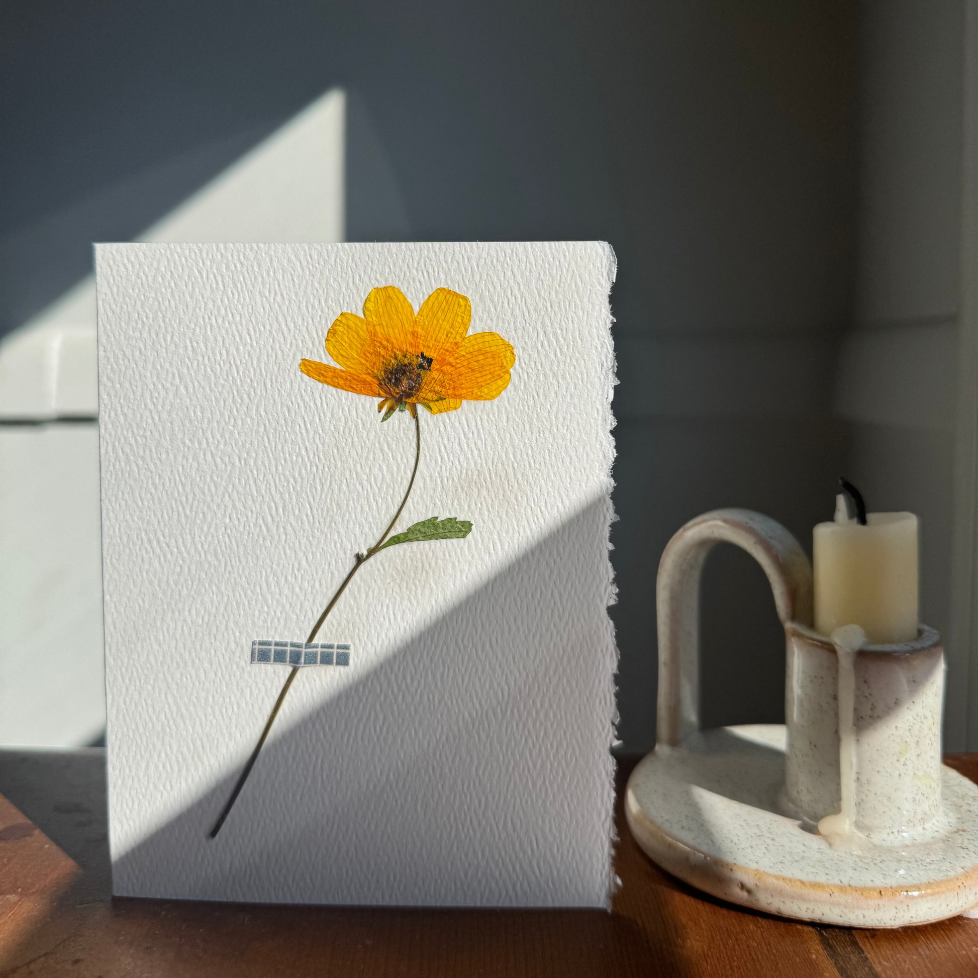 White greeting card with a pressed yellow flower on a wooden surface with a candle holder in the background.